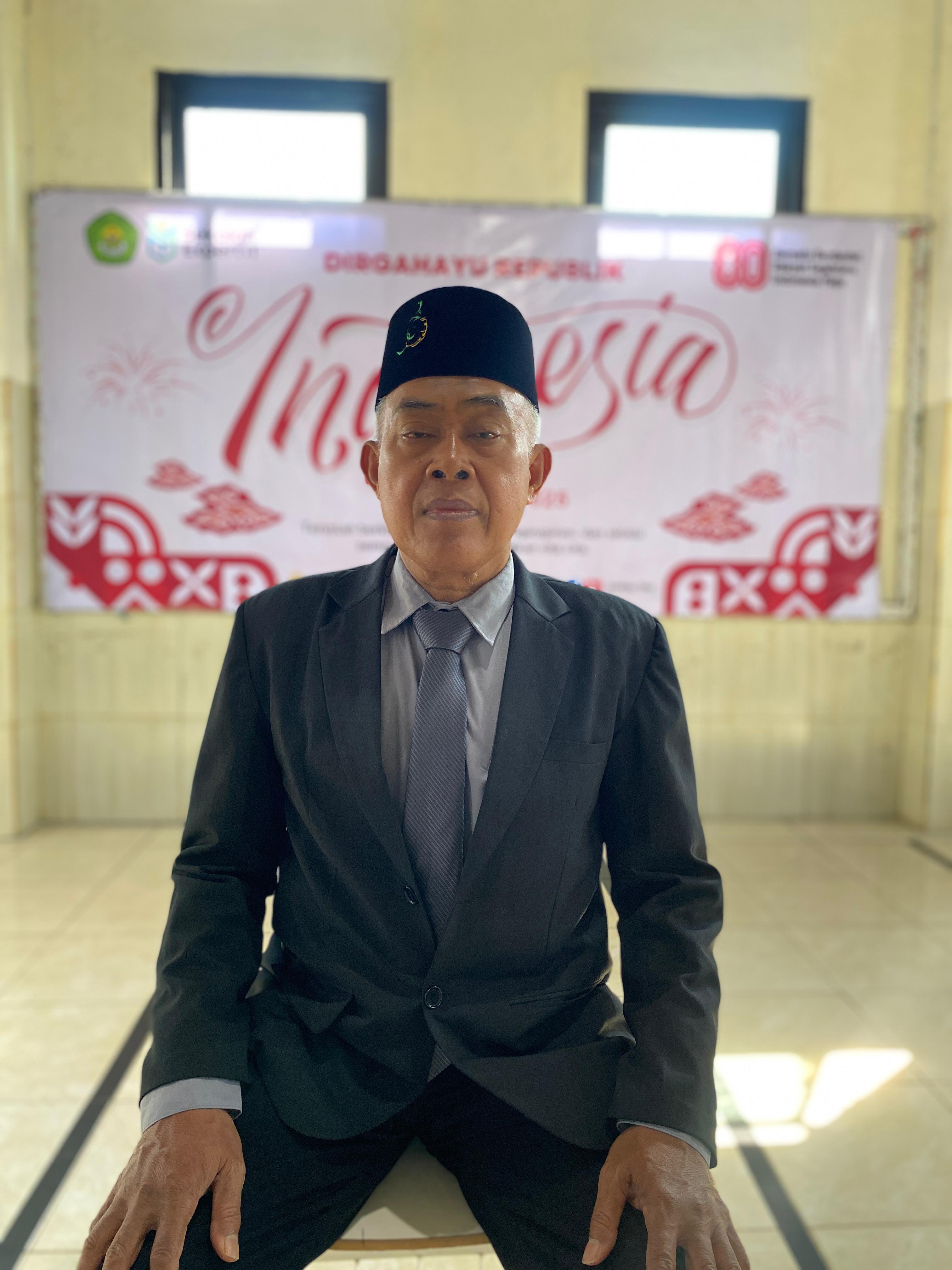 Kepala Sekolah Drs. H. M. Roji, M.Pd. memakai jas hitam dan peci hitam sedang tersenyum pada latar merah putih bertulisan Muhammadiyah